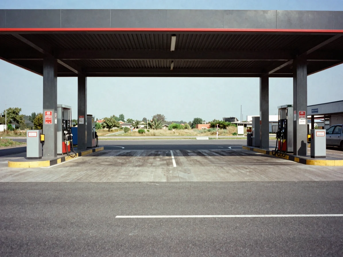 Editorial commercial photograph of a station forecourt approach seen from the roadway, clear daylight, steel and charcoa
